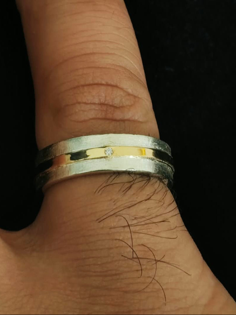 A close-up of a person's finger wearing a silver band ring with a polished gold inlay, set against a solid black background.