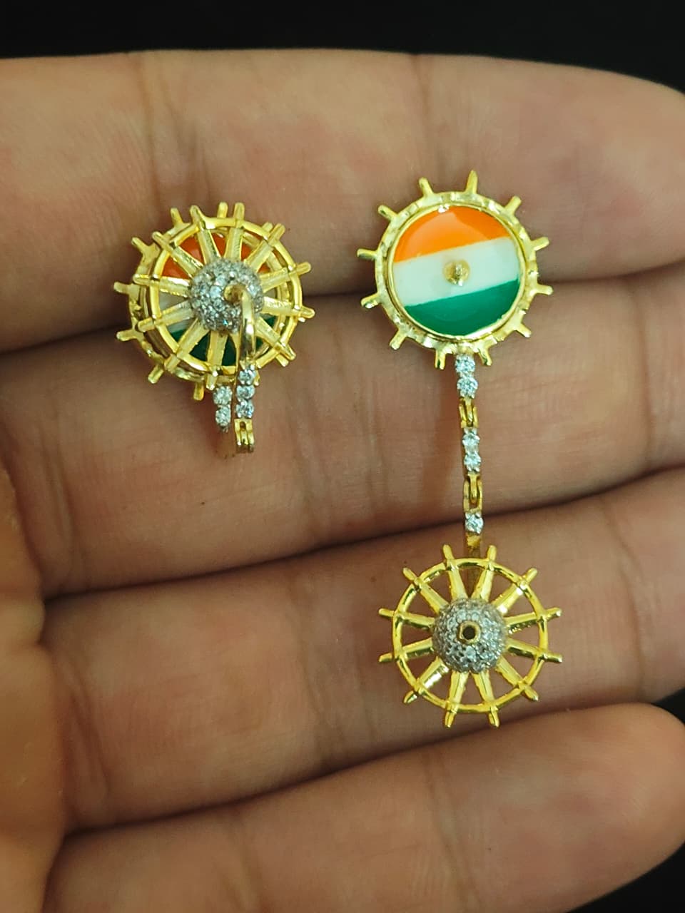 A pair of gold-toned earrings displayed on a person's hand. One earring features a central oval inlay with the Indian flag (saffron, white, and green), and the other is a matching gold wheel design with a central stone cluster.