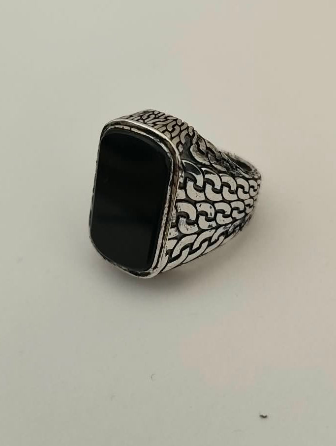 A silver signet ring with an engraved patterned band and a black rectangular cabochon stone, resting on a plain off-white surface.