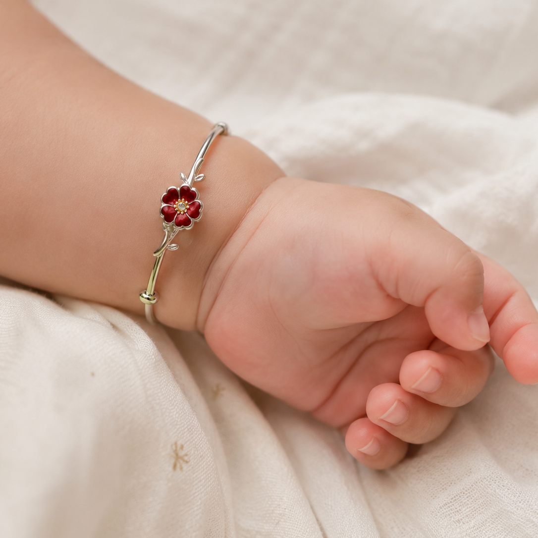 Baby's hand wearing a silver bracelet with a red flower charm on a white fabric background
