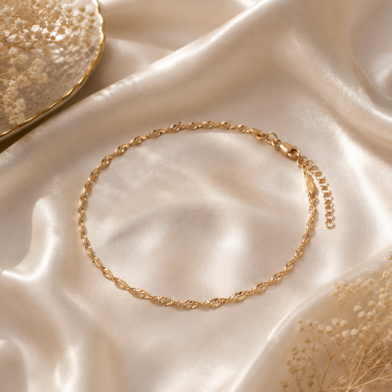 Gold chain anklet on a textured beige surface with dried flowers.