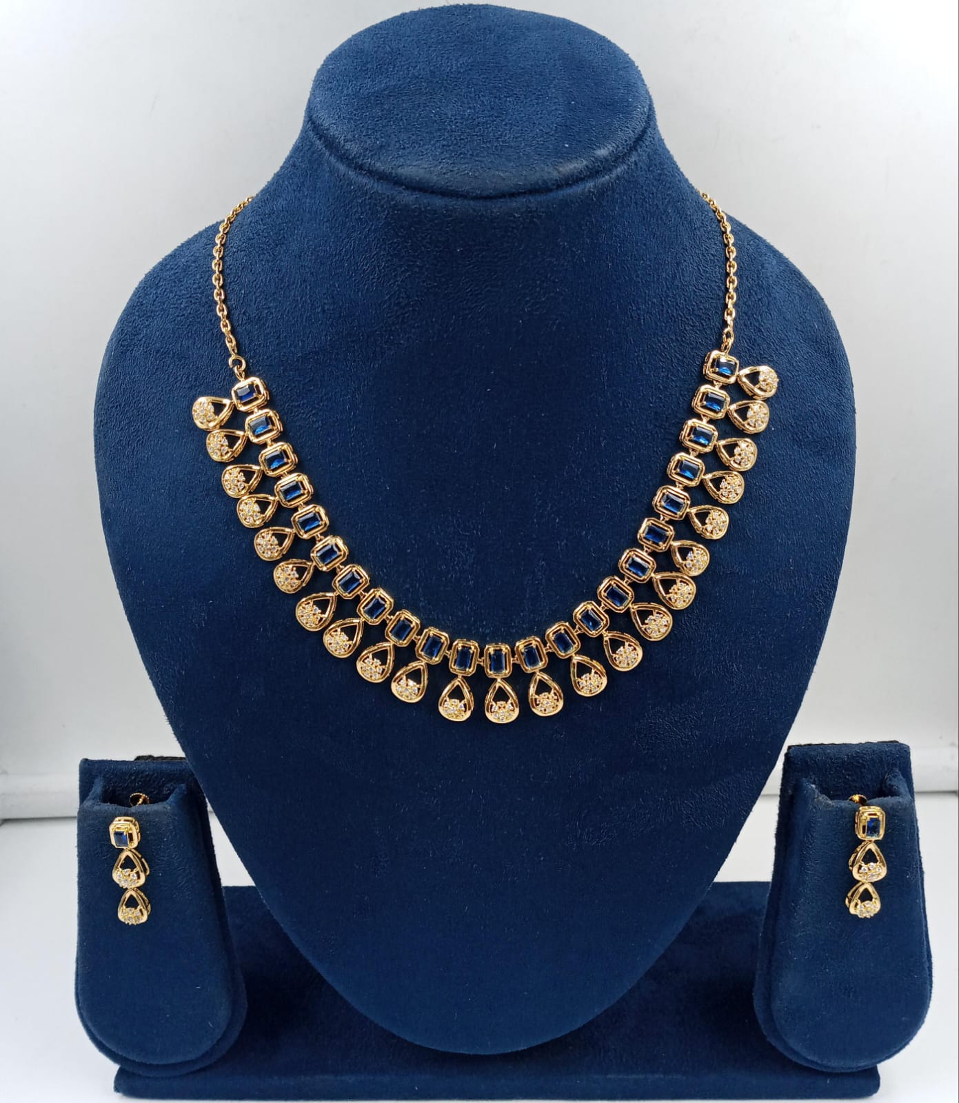Royal Blue Sapphire Necklace Set by Rivansh featuring a gold-toned custom sterling silver crescent necklace and matching drop earrings meticulously arranged with square-cut sapphire blue stones and brilliant pave accent halos on a premium velvet bust display.