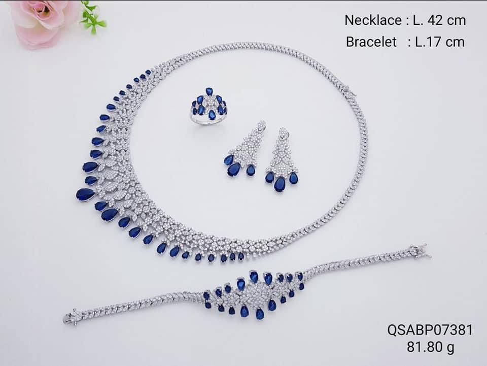 Sapphire Blue Necklace Set in rhodium-finished 925 silver featuring a double crescent layout of graduated teardrop-cut blue composite gemstones with matching chandelier earrings, ring, and line bracelet by Rivansh.