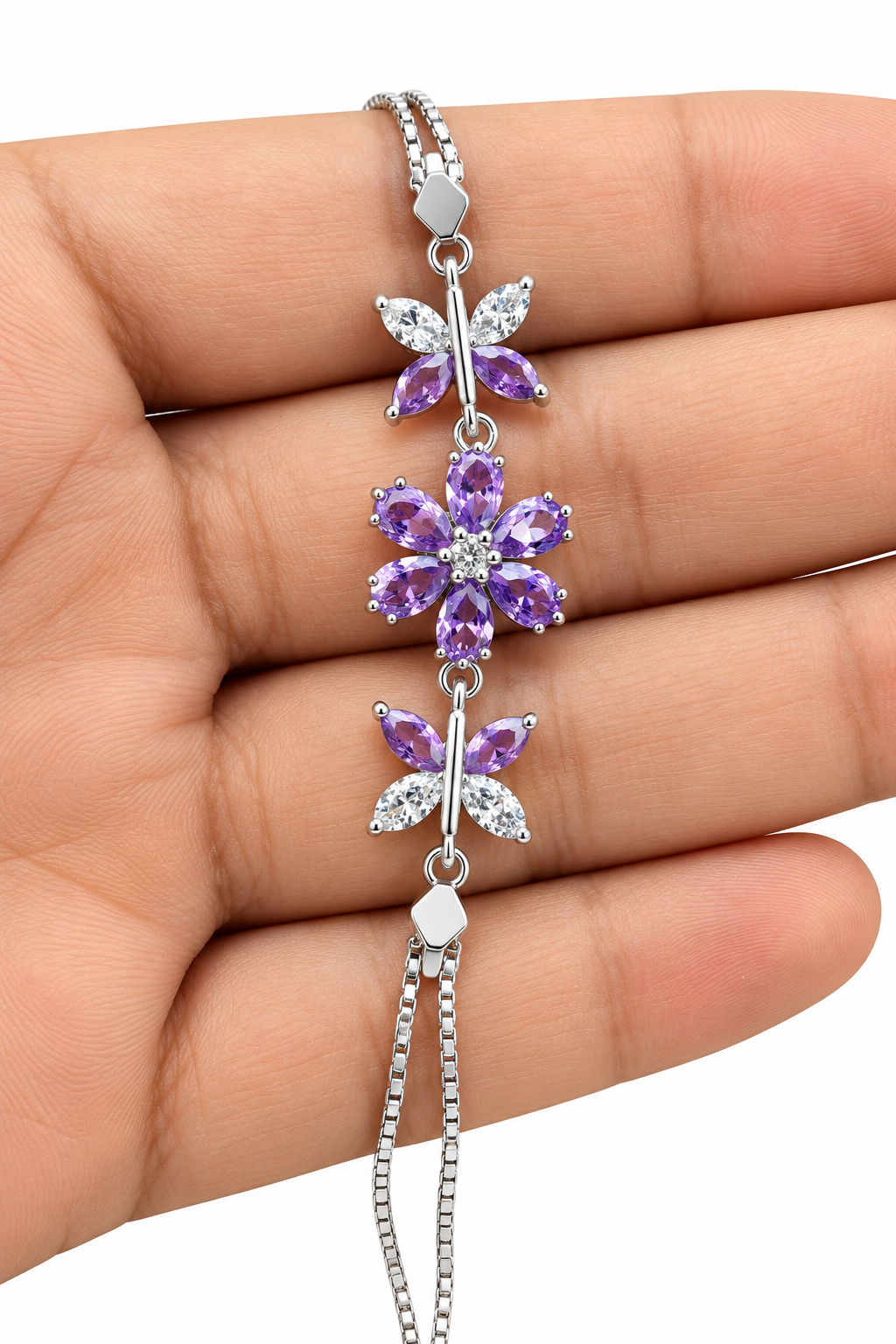 A silver-toned bracelet with three purple flower and butterfly charms, held in a person's hand against a white background.