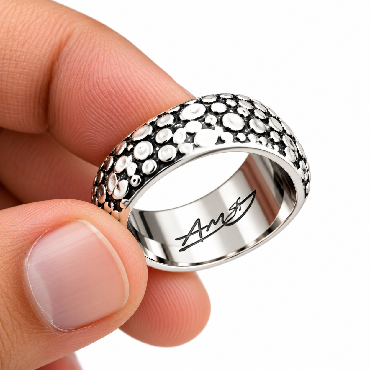 A close-up of a hand holding a silver band ring decorated with small round clear stones on the exterior and an engraved script logo on the interior, against a plain white background.